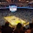 An indoor basketball arena filled with spectators. The court features a central logo and is surrounded by partially occupied tiered seating. A large scoreboard hangs above the center of the court, displaying the game score and time. Ambient lighting highlights the players on the court and the vibrant atmosphere of a live sports event.