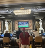 Several people are seated at a luxurious bar, with a large TV screen showing a sports game. The bar is elegantly decorated with ornate details and sculptures. The ceiling has a large, rectangular, crystal chandelier.